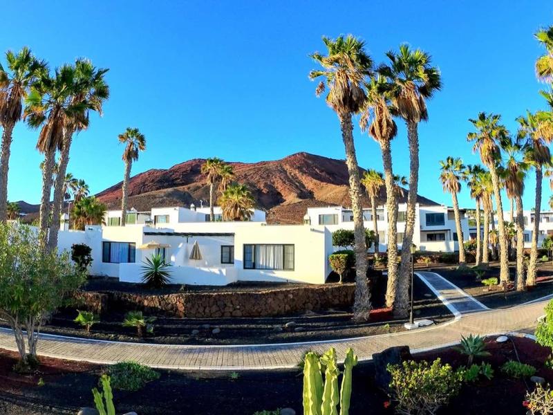 White buildings and palm trees in front of a dry hill under clear blue sky.