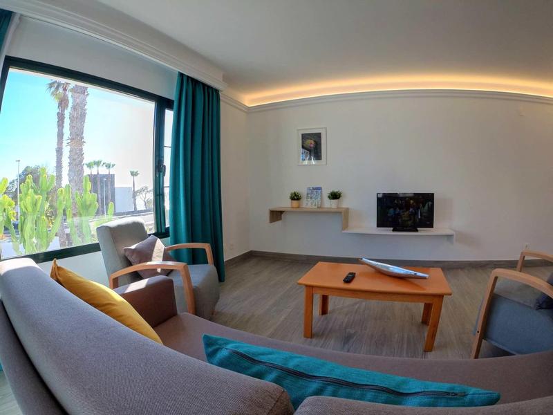 Modern living room with sofa, chairs, TV, and large window offering a view of palm trees.