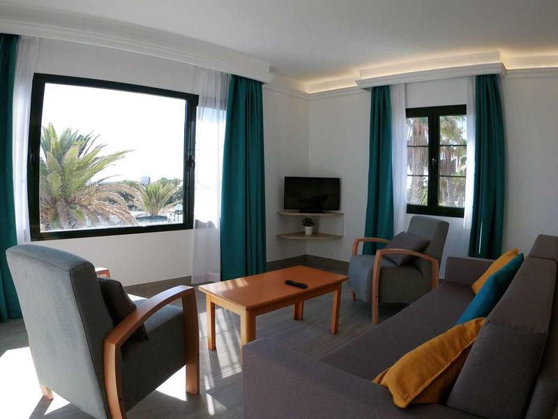 Bright, modern living room with sofa, armchairs, TV, and view of palm trees.