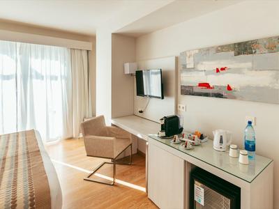 Helles Hotelzimmer mit Bett, Stuhl, Fernseher und großem Fenster mit Vorhängen.