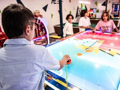 Junge spielt an einem bunten Air-Hockey-Tisch in einer belebten Spielhalle mit mehreren Spielern.