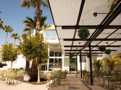 Offener Sitzbereich mit Metallstühlen und Tischen, Palmen und moderner Glasfassade im Hintergrund.