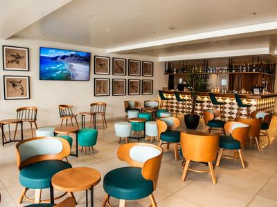 Moderne Bar mit Holzstühlen, grünen Sesseln, Tischen und großer Bildschirm an weißer Wand.