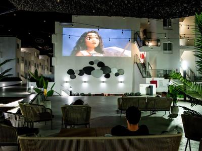 Freiluftkino bei Nacht mit großer Leinwand, Sitzgelegenheiten und grünen Pflanzen.