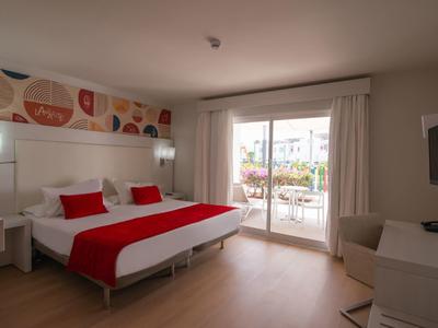 Modernes Gästezimmer mit Doppelbett, roten Akzenten, Balkon und heller Holzbodenoptik.