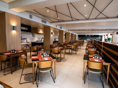 Moderner, heller Restaurantraum mit Holzmöbeln, vielen Tischen und Stühlen, weißen Wänden und Deckenspotlights.