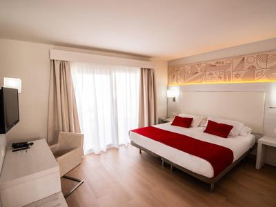 Helles Hotelzimmer mit Doppelbett, roter Bettdecke, Fernseher, Sessel und großem Fenster mit Vorhängen.