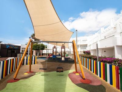 Kinderspielplatz mit buntem Zaun, Schaukeln, Sonnensegel und weichem Bodenbelag.