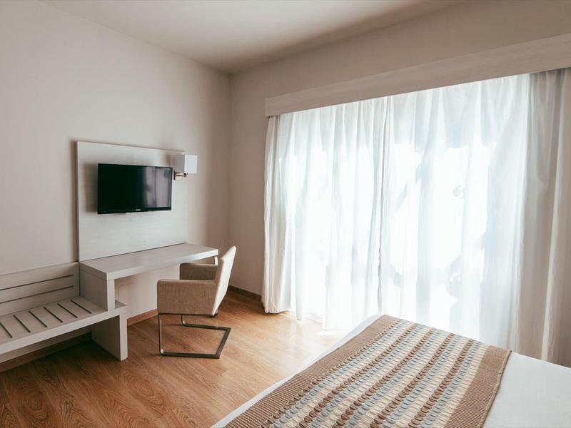 Helles, schlicht eingerichtetes Schlafzimmer mit Fensterfront, Bett, Stuhl und Wandfernseher.