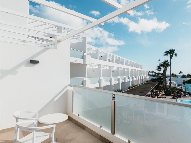Balkon mit weißen Möbeln, Glasgeländer, Blick auf Palmen und Meer unter blauem Himmel mit Wolken.