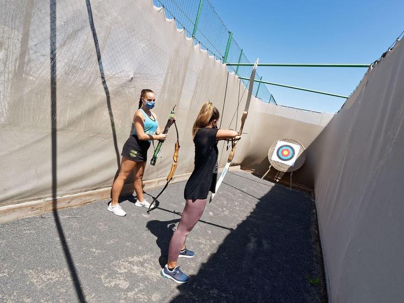 Zwei Frauen üben Bogenschießen in einem sonnigen, eingezäunten Bereich mit Zielscheibe.