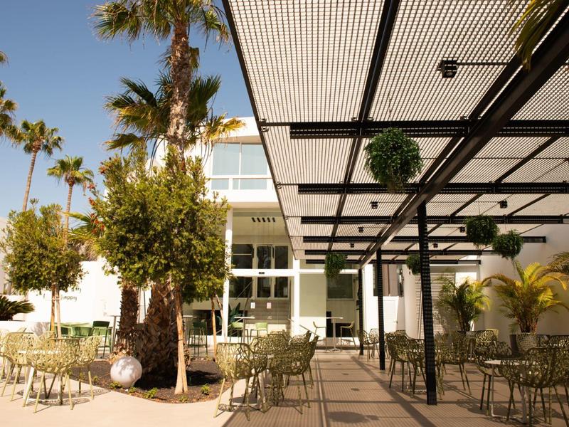 Offener Sitzbereich mit Metallstühlen und Tischen, Palmen und moderner Glasfassade im Hintergrund.