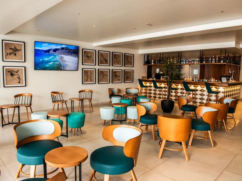 Moderne Bar mit Holzstühlen, grünen Sesseln, Tischen und großer Bildschirm an weißer Wand.