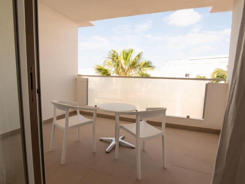 Balkon mit zwei weißen Stühlen und kleinem runden Tisch, Blick auf Palmen und blauen Himmel.