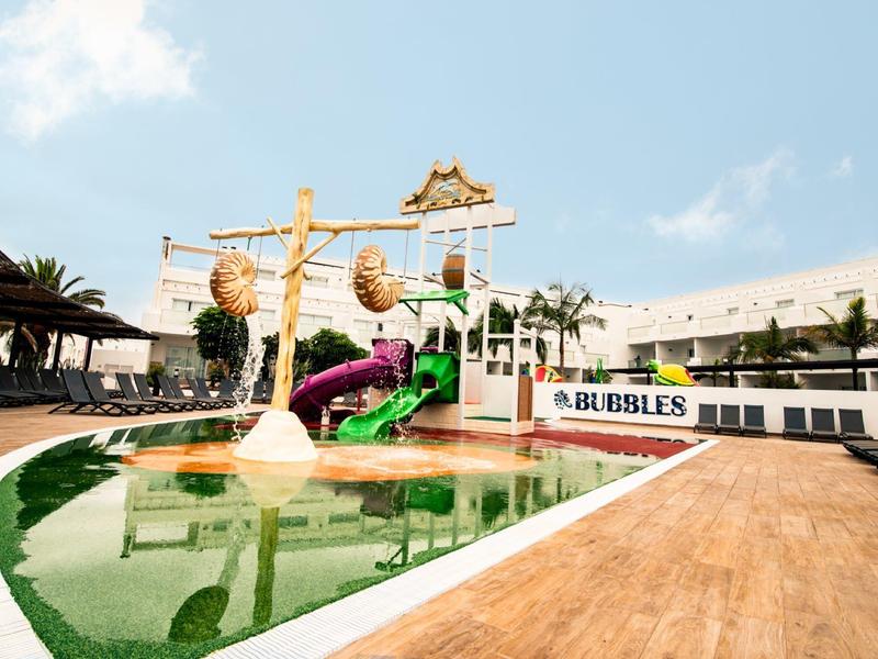 Ein kinderfreundlicher Wasserspielplatz mit Rutschen, Wasserkanonen und Palmen unter blauem Himmel.