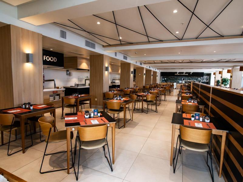 Moderner, heller Restaurantraum mit Holzmöbeln, vielen Tischen und Stühlen, weißen Wänden und Deckenspotlights.