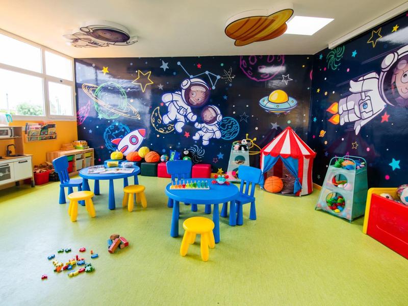 Bunter Kindergartenraum mit Weltraumwandbild, Tischen, Stühlen, Spielzeug und Zelt.