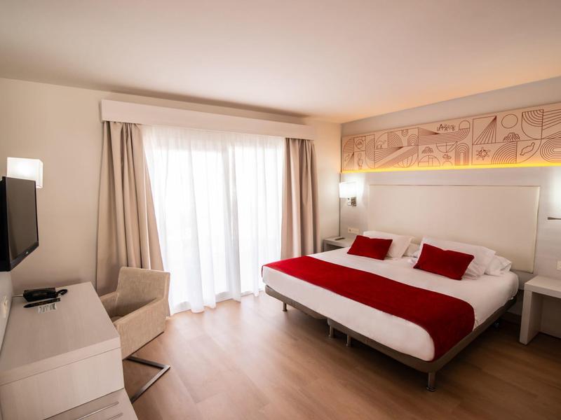 Helles Hotelzimmer mit Doppelbett, roter Bettdecke, Fernseher, Sessel und großem Fenster mit Vorhängen.