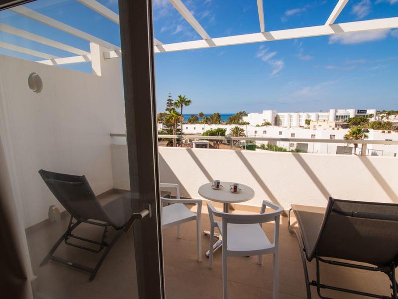 Balkon mit Stühlen, kleinem Tisch, Glaswand und Blick auf weiße Häuser und blauen Himmel.