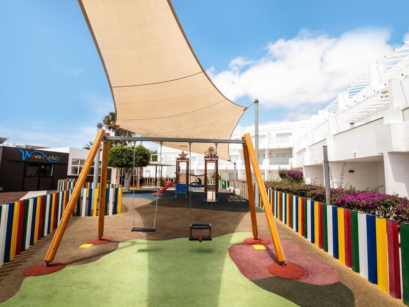 Kinderspielplatz mit buntem Zaun, Schaukeln, Sonnensegel und weichem Bodenbelag.