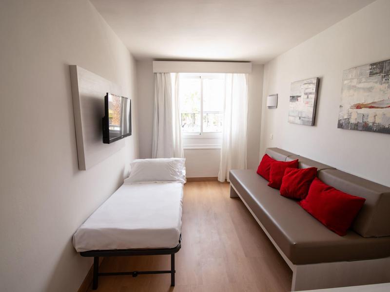 Kleiner Raum mit zwei Sofabetten, weißen Wänden, Holzfußboden und Fenster mit weißen Vorhängen.