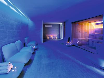 Moderner Wellnessraum mit blauer Beleuchtung, Sofas links, großer Glaswand rechts und Kerzen am Boden.