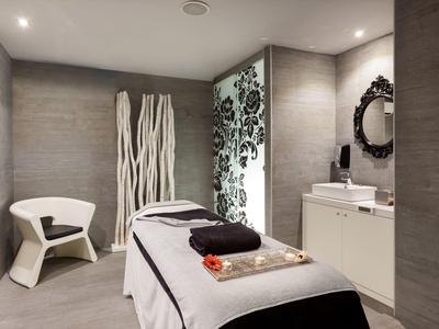 Moderne Wellnessmassage mit grauen Wänden, weißem Stuhl, dekorativen Stäben und beleuchtetem Spiegel.