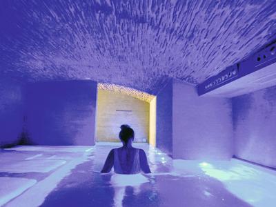 Person sitzt im beleuchteten Wasserpool in einem dunklen Raum mit blauen und violetten Farbtönen.