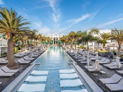 Großer rechteckiger Pool mit Palmen, Sonnenliegen und weißem Hotelgebäude bei blauem Himmel.