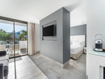 Moderne Wohnung mit grauem Fliesenboden, TV an Trennwand, Schlafzimmer und Balkon mit Glas-Schiebetür.