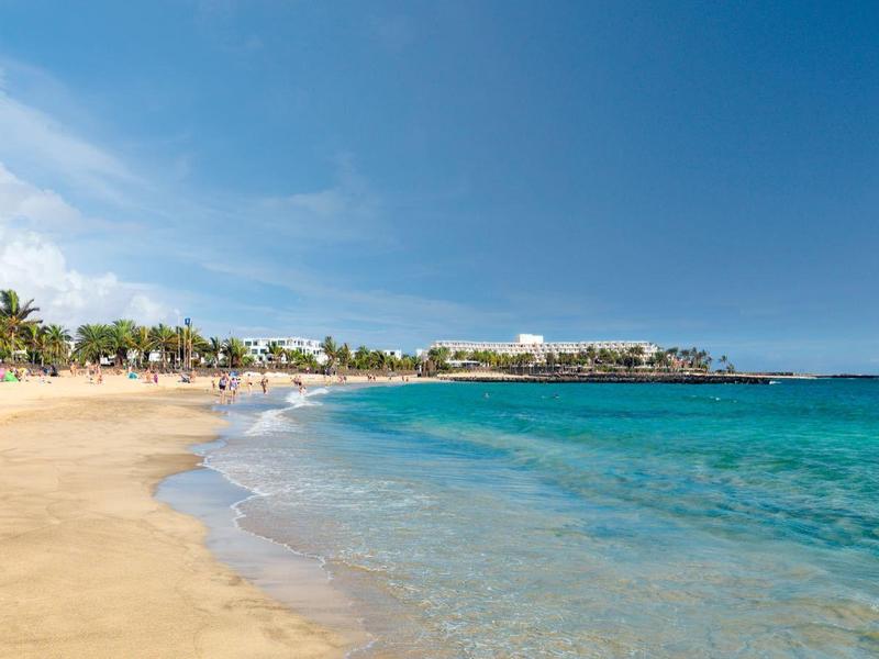 Spiaggia di sabbia dorata con acqua turchese e palme sotto un cielo parzialmente nuvoloso.