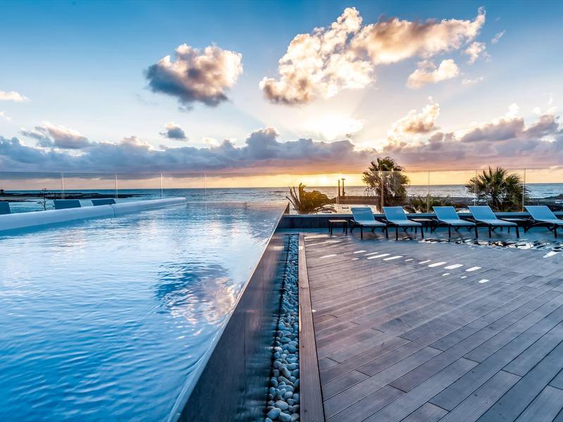 Piscina a sfioro con vista sul mare al tramonto, con sdraio sulla terrazza.