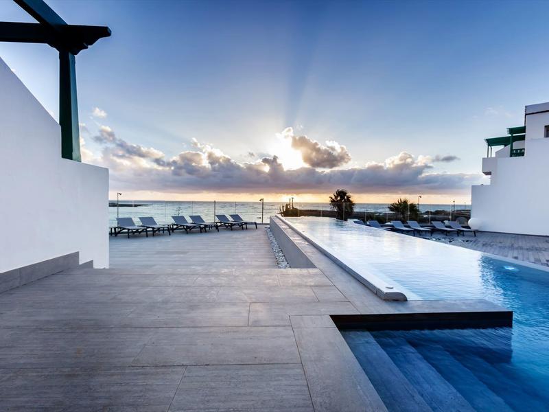 Piscina moderna con lettini di fronte al tramonto, cielo azzurro e mare all'orizzonte.