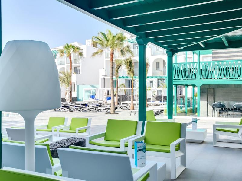 Terrazza di hotel moderna con sedute verdi e vista su piscina e palme soleggiate.