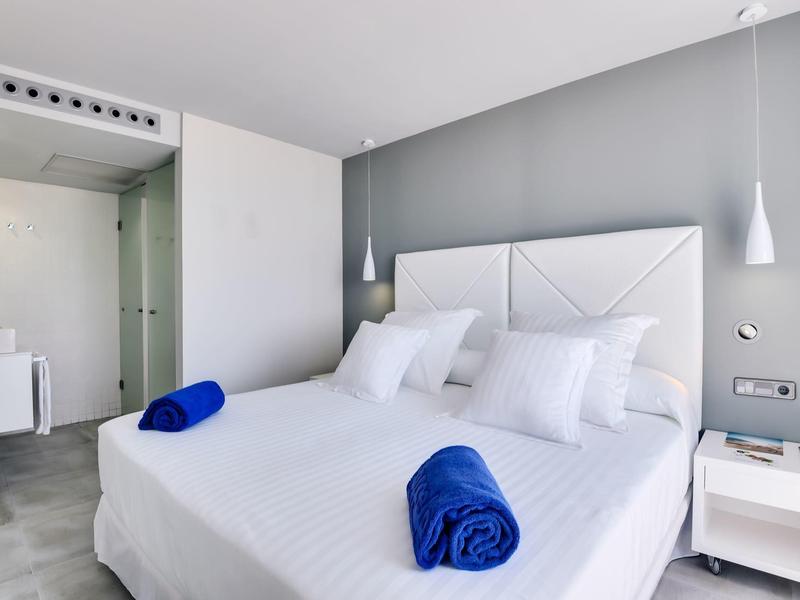 Camera d'hotel moderna con letto matrimoniale bianco, pareti grigie e asciugamani blu arrotolati.