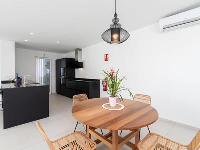 Modern dining area with round wooden table and plants beside an open kitchen with black furnishings.
