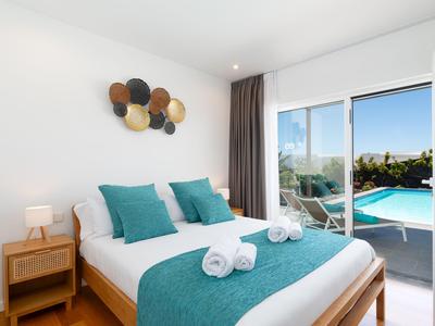 Helles Schlafzimmer mit Doppelbett, türkisfarbenen Kissen und Blick auf den Pool durch die Schiebetür.