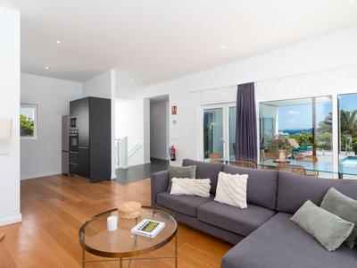 Helles Wohnzimmer mit großem grauem Sofa, Glastisch und Blick auf Terrasse mit Pool und Palmen.