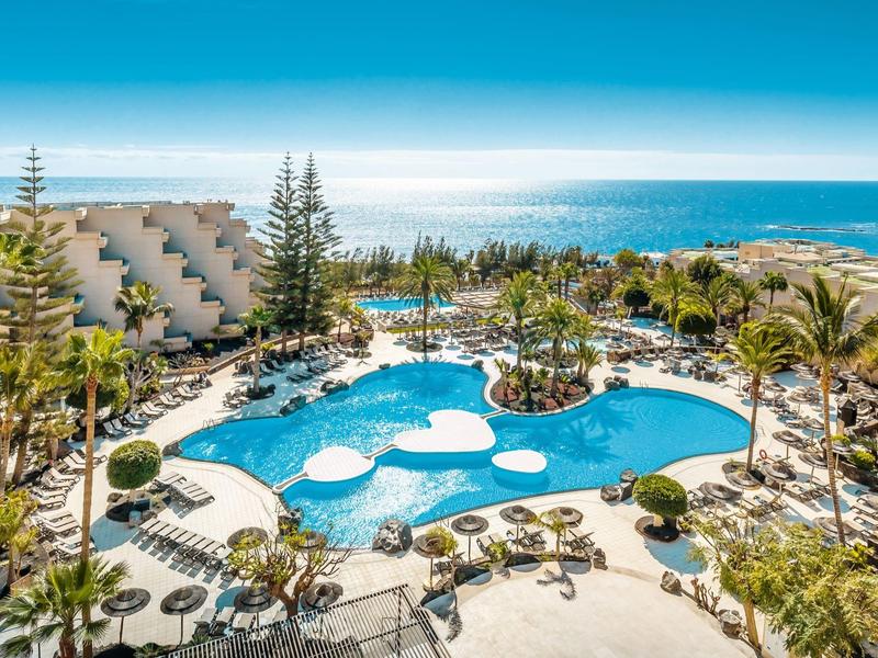 Large hotel pool with palm trees, lounge chairs, and sea view under blue sky.