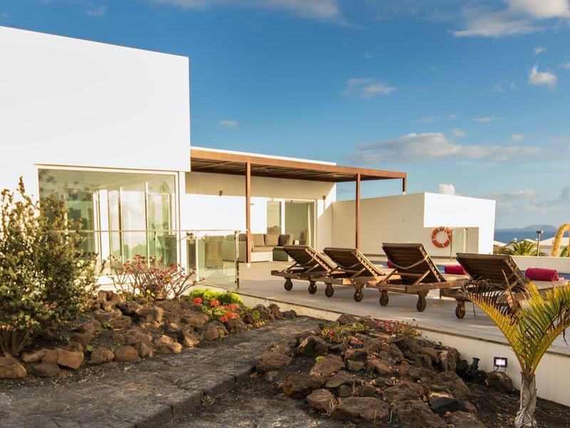 Modern holiday home with pool and sun loungers under clear sky overlooking the sea.