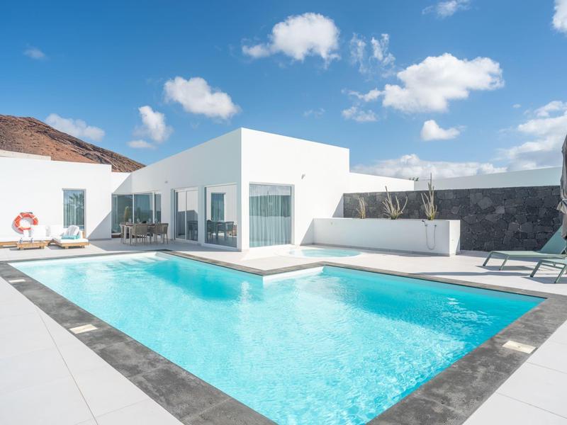 Modern white bungalow with clear pool and mountain backdrop under sunny weather.