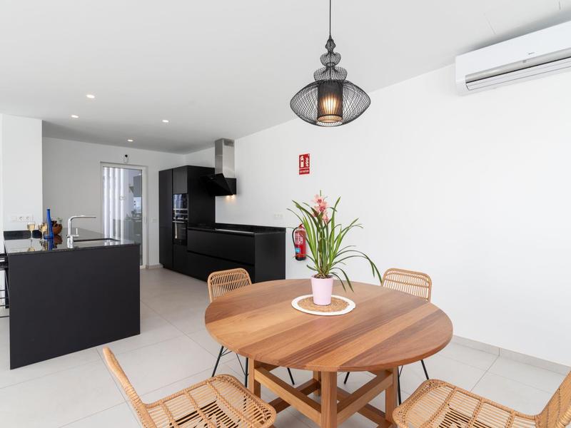 Modern dining area with round wooden table and plants beside an open kitchen with black furnishings.