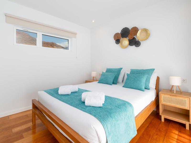 Modern bedroom with wooden bed, turquoise pillows and blanket, wooden floor, and nightstand with lamp.
