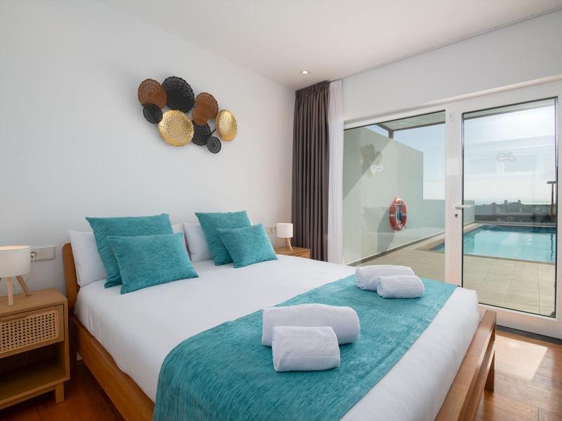 Modern hotel room with double bed, turquoise pillows and blanket, and access to a balcony with pool view.