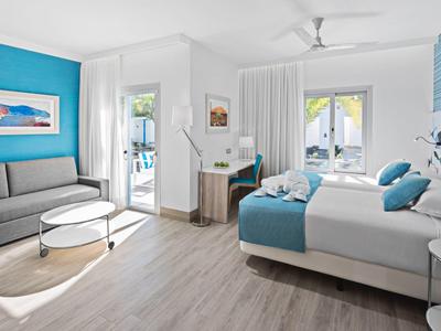 Helles, modernes Hotelzimmer mit Doppelbett, grauem Sofa, Schreibtisch und blauen Akzenten.
