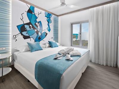 Modernes Schlafzimmer mit weißem Bett, blauer Bettdecke, abstraktem Wandbild und großen Fenstern.