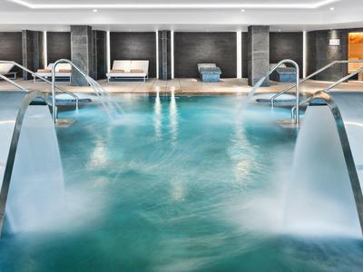 Innen-Schwimmbad mit Wasserfällen, Edelstahlgeländern und entspannenden Liegen im Hintergrund.