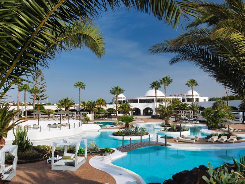 Área de piscina del hotel con palmeras, edificio blanco y cielo azul de fondo.