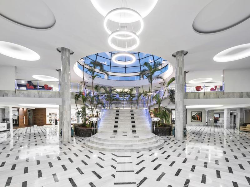 Vestíbulo moderno de hotel con suelo de mármol blanco, escalera central y aberturas circulares en el techo.