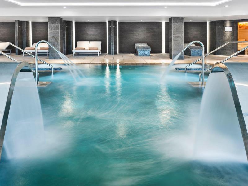 Zona de piscina cubierta moderna con cascadas, tumbonas y ambiente relajado en el hotel.